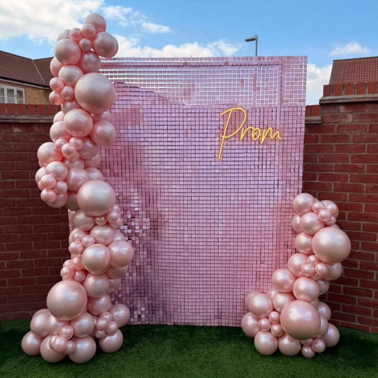 A pink party backdrop setup featuring a dusty pink sequin wall with a yellow neon 'Prom' sign, flanked by two tall balloon garlands and a smaller balloon cluster on the right, set outdoors on artificial grass against a brick wall.